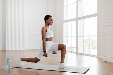 Black woman doing a lunge workout holding dumbbells in a while apartment