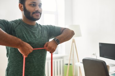 For strong arms. Cropped shot of young active man looking away, exercising with resistance band during morning workout at home. Sport, healthy lifestyle